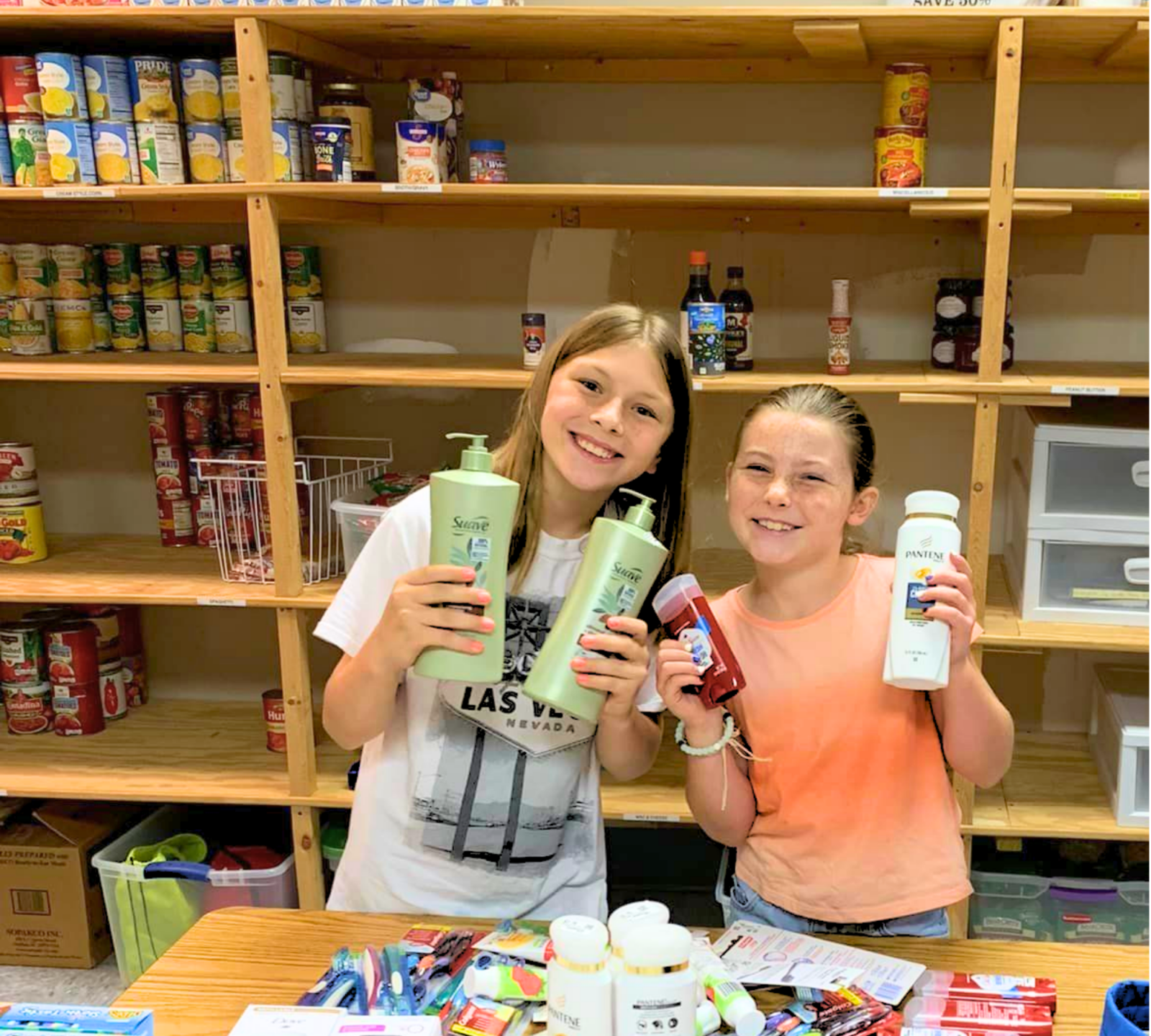 Two young girls smile and hold up shampoo and body wash as they help stock the Helping Hands pantry.